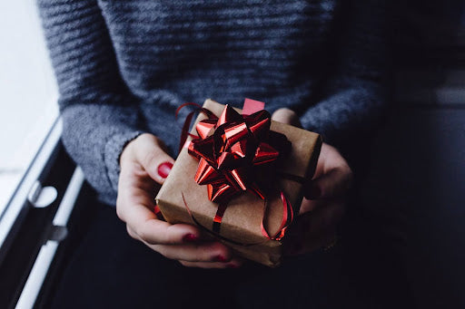 Photo of a woman’s hands, holding a small box-shaped gift wrapped in brown paper, and a red satin bow.