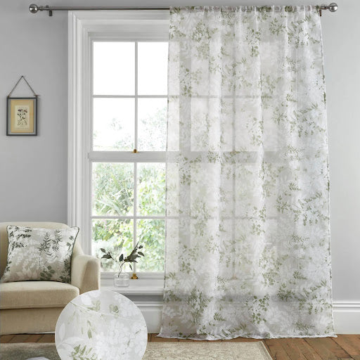 Sheer white curtains with a delicate green floral pattern hang from a metal rod in a room with soft natural light. The interior includes a cream armchair and a framed picture on a grey wall. 