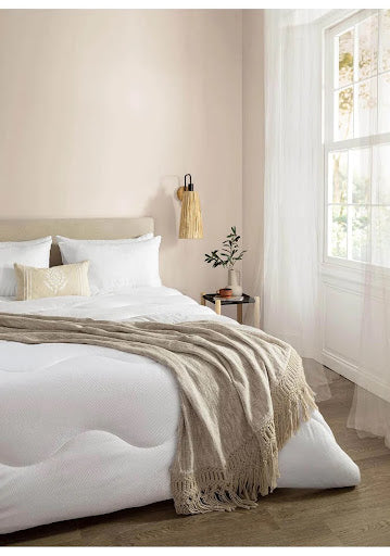  A bedroom with a bed with white duvet and a textured beige throw, and a window dressed with sheer curtains that offer a view of the outdoors.