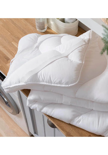A stack of white, quilted mattress enhancers by Shaws is placed atop a wooden surface, with a plush appearance indicative of added comfort and support