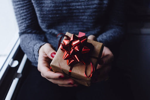 Photo of a woman’s hands, holding a small box-shaped gift wrapped in brown paper, and a red satin bow. 