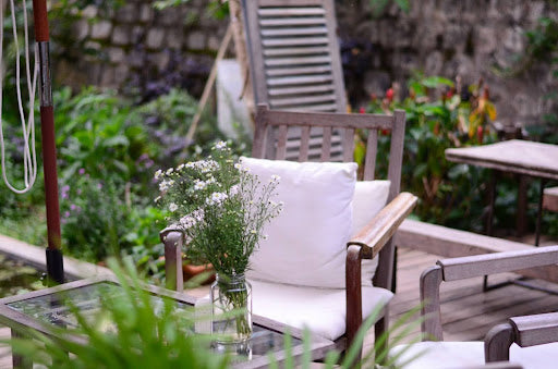 A cosy outdoor seating area with wooden chairs and white cushions featuring a glass table with a jar of delicate white flowers, surrounded by lush green plants and a stone wall in the background.