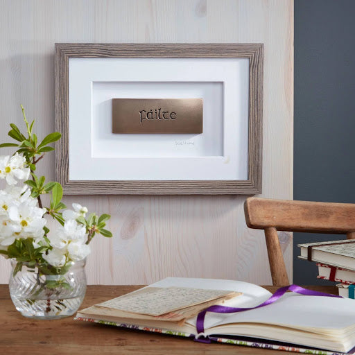 Framed artwork with the word “Failte” (Welcome) in the centre of the frame. It is set behind a table and chair, with a vase of flowers and book on the table. 