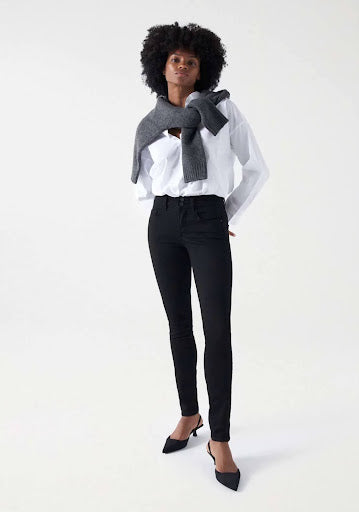 A woman wearing black skinny jeans, a white shirt, and a knitted sweater tied around her shoulders.
